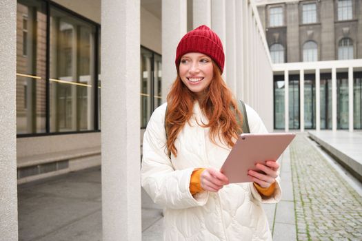 Stylish ginger girl, tourist walks with digital tablet around city, woman connects to iternet on her gadget, looking up information, texting message.