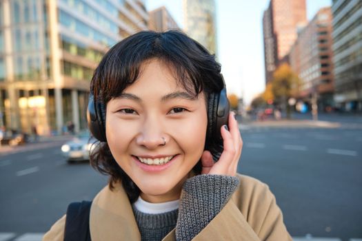 Portrait of young smiling korean girl, walking along city centre, listening music in headphones and holding mobile phone. Copy space