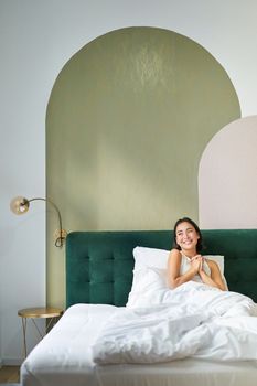Vertical shot of beautiful asian girl lying in bedroom with sophisticated interrior, smiling and looking happy in morning, spending time in bed.