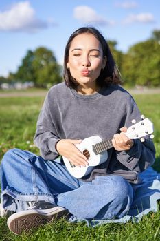 Positive hipster asian girl, playing ukulele and making faces, singing and feeling happy.