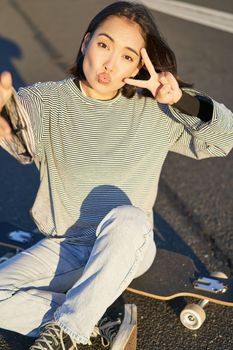 Selfie of asian girl sitting on skateboard, taking photo on smartphone, smiling and showing peace v-sign.