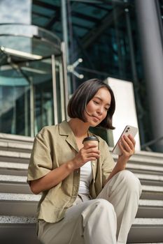 Beautiful smiling asian girl, drinking coffee, using mobile phone and sits on stairs outside. Young woman video chat outdoors.