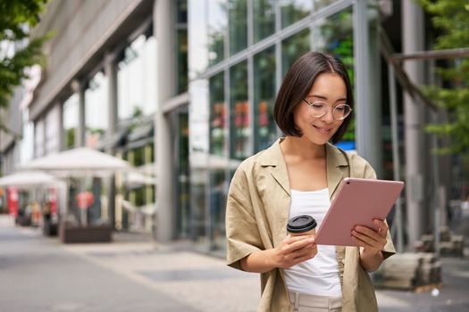 Portrait of smiling young woman walking in city with tablet, drinking takeaway coffee, going down the street with happy expression.