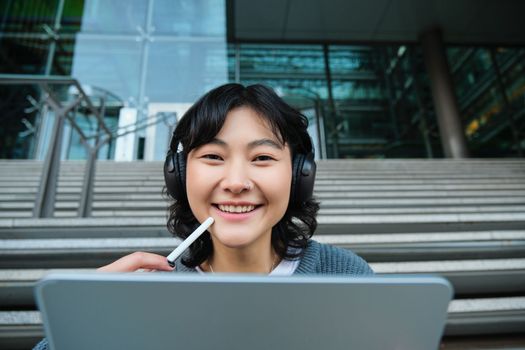 Close up of smiling asian girl feels inspired while draws on digital tablet, makes graphic design project, pencils user interface assignment, sits on stairs near campus.