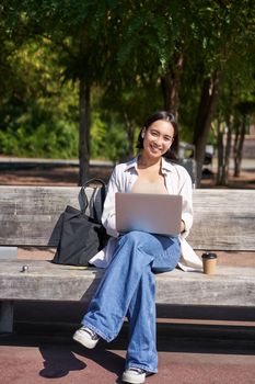 Portrait of asian woman sitting with laptop on bench in park, listening music with wireless headphones, doing homework, working on remote.