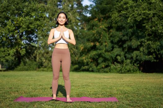 Young mindful woman, meditating on fresh air, doing yoga exercises in park, standing on rubber mat in sportswear.