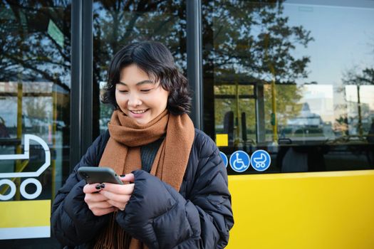 Young beautiful woman standing on bus stop, texting message on smartphone, holding mobile phone, checking her schedule, buying ticket online, wearing winter clothes.