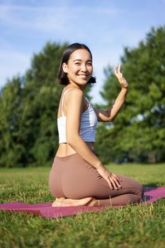 Portrait of asian girl on fitness class in park, sitting on rubber mat and wave hand at camera, say hello, meditating and practice yoga.