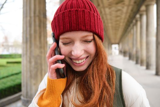 Portrait of redhead european girl in red hat, makes a phone call, walks in city and talks to friend on smartphone. Copy space