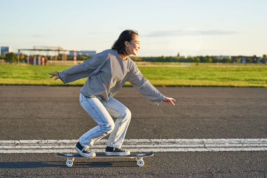 Happy skater girl riding her skateboard and having fun on empty street. Smiling woman enjoying cruiser ride on sunny road.