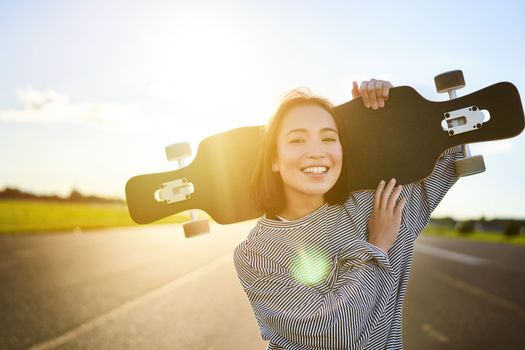 Happy skater girl walking towards the camera, sunbeams shining at lense. Young woman skating on cruiser, holding longboard on shoulders.