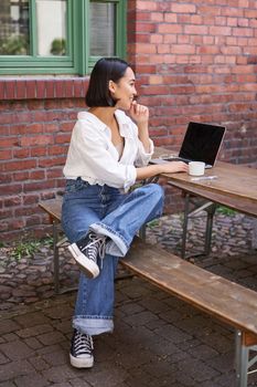 Stylish young businesswoman, asian girl with laptop, sitting in outdoor cafe with cup of coffee and working, using computer.