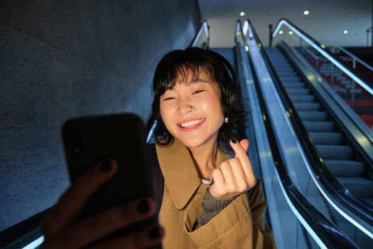 Stylish smiling asian girl, taking selfie on mobile phone while riding escalator, going down to metro station, showing finger heart and posing for photo.