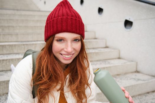 Smiling traveler, redhead girl tourist sits on stairs with flask, drinks hot coffee from thermos while travelling and sightseeing around foreign city, sits on stairs and rests.