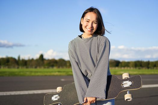 Portrait of beautiful asian girl skating on longboard, crusing with skateboard on empty road, enjoying freetime on fresh air.