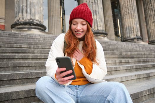 Young smiling woman with long red hair, sits near building on stairs with smartphone, using mobile application, sending message.