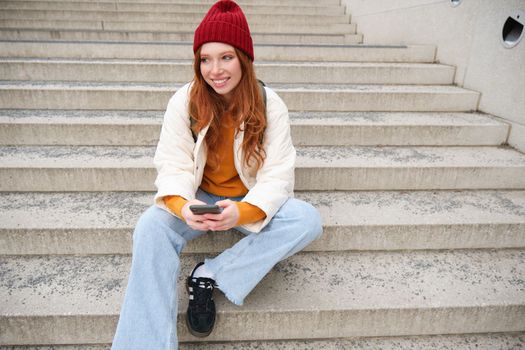 Hipster ginger girl, redhead woman sits on stairs with smartphone, waits for someone and messages on social media on mobile phone app. People and technology concept