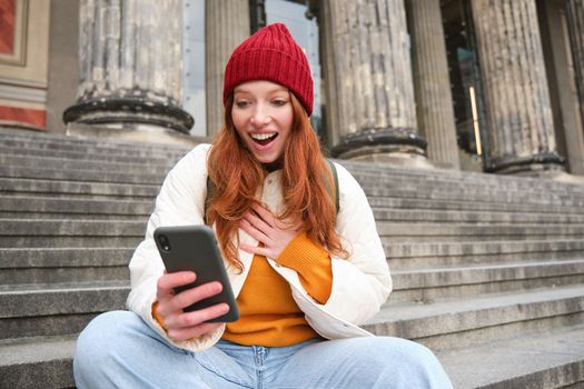 Redhead female tourist talks on mobile phone, video chatting with friends, says hi and waves hand at smartphone camera.