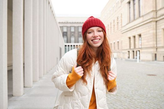 Female tourist in red hat with backpack, sightseeing, explores historical landmarks on her trip around europe, smiling and posing on street.