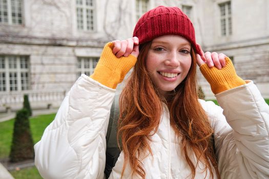 Young smiling redhead woman walking in beautiful city attractions, wearing backpack, red hat and coat, looking around with happy face.