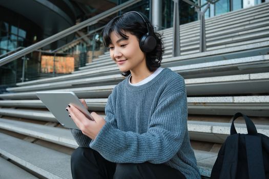 Smiling chinese student sits on stairs with tablet, draws digital art, graphic design project for freelance job, listens music in headphones and smiles.