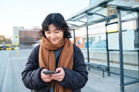 Happy smiling korean girl, using mobile phone, standing on bus stop with smartphone, looking at departure schedule on application, posing in winter clothes.