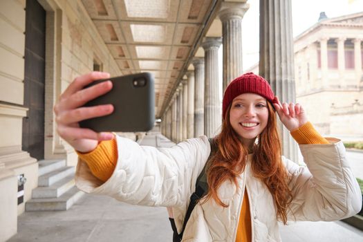 Cute young redhead woman takes selfie on street with mobile phone, makes a photo of herself with smartphone app on street.