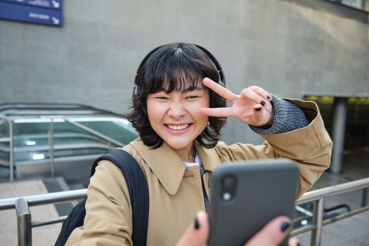 Beautiful korean girl in headphones, takes selfie on her smartphone, stands on street with mobile phone, records video, listens music and travels around city.