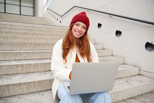 Smiling redhead girl, student sits on stairs outdoors and uses laptop, connects to public wifi in city and works on project, uses internet on computer.