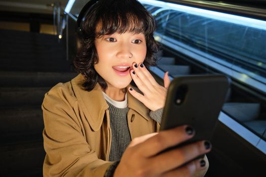 Close up portrait of korean girl looks surprised at mobile phone, reads something amazing on smarpthone screen, watching video in headphones, sits on staircase.