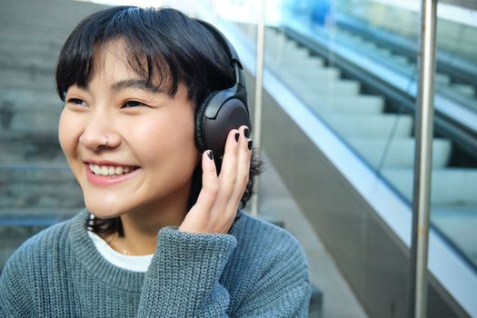 Portrait of smiling korean girl in headphones, uses smartphone and sits on stairs in mall, watches video on mobile phone. Copy space