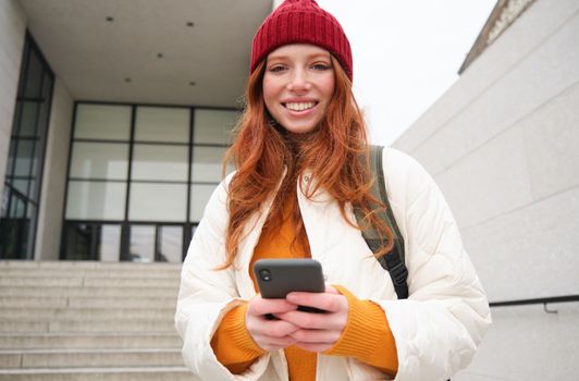 Happy girl student in red hat, holds smartphone, tourist looks at map app on her phone, explores sightseeing, texts message, looks for couchsurfing, rents place to stay online.