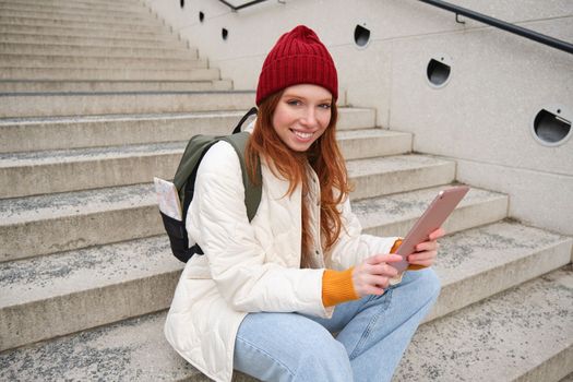 Happy stylish redhead girl, student in red hat, holds digital tablet, uses social media app, searches something online, connects to wifi.