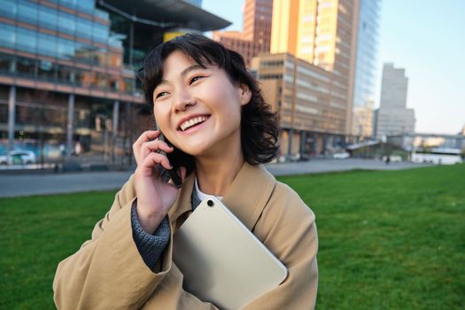 University lifestyle. Young asian student, girl with tablet and notebook, talks on mobile phone while walks from campus on street.