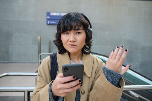 Portrait of asian girl in headphones, looks complicated at smartphone screen, puzzled by text message or notification, stands on street and shrugs.