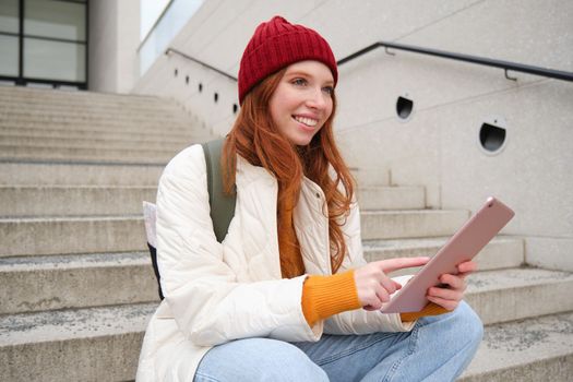 Happy stylish redhead girl, student in red hat, holds digital tablet, uses social media app, searches something online, connects to wifi.