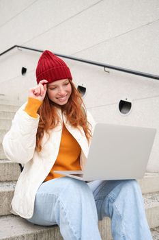 Smiling redhead girl, student sits on stairs outdoors and uses laptop, connects to public wifi in city and works on project, uses internet on computer.