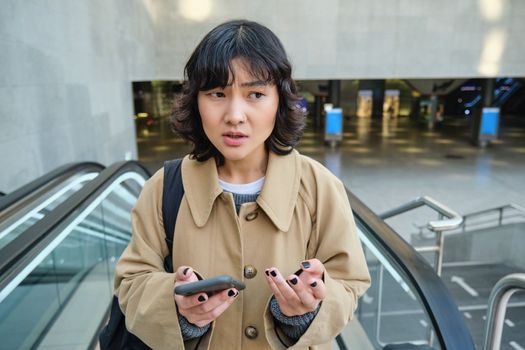 People in city. Portrait of korean girl standing on escalator, looks confused after reading text message on mobile phone.