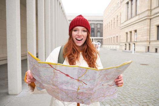 Beautiful redhead woman, tourist with city map, explores sightseeing historical landmark, walking around old town, smiling happily.