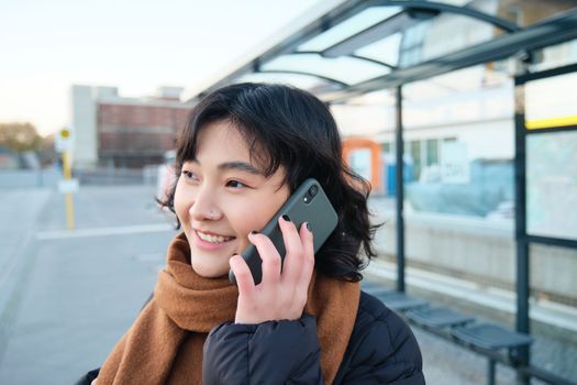 Close up of young asian woman talks on mobile phone, has conversation over telephone, waits for bus on public transport stop.