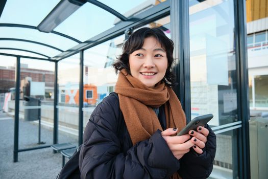 Beautiful young woman, standing on bus stop, waiting for her public transport, using mobile phone, using smartphone application.