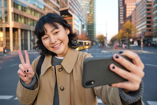Happy asian girl, tourist takes photo in city centre, shows peace sign at smartphone camera, makes selfie during sightseeing with smiling joyful face.