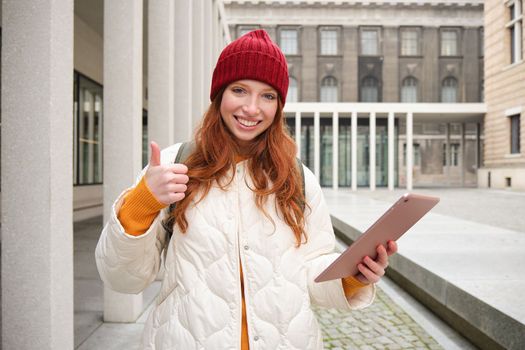 Stylish ginger girl, tourist walks with digital tablet around city, woman connects to iternet on her gadget, looking up information, texting message.