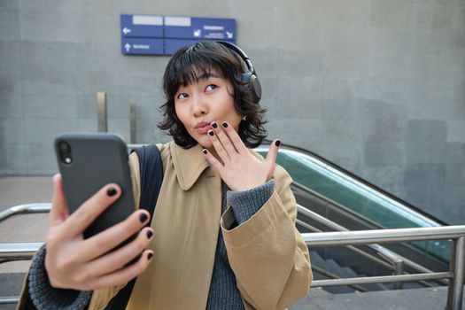 Cute and funny korean girl in headphones, posing for selfie, takes photo on smartphone and looks silly, stands on street in city.