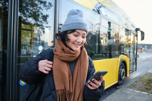 Image of girl student waiting for public transport, checks schedule on smartphone app, stands near city bus. Urban lifestyle concept