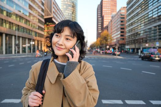 Smiling asian girl makes a phone call, stands on an empty street, calling someone on telephone, waiting for friend in city, going to a meeting.