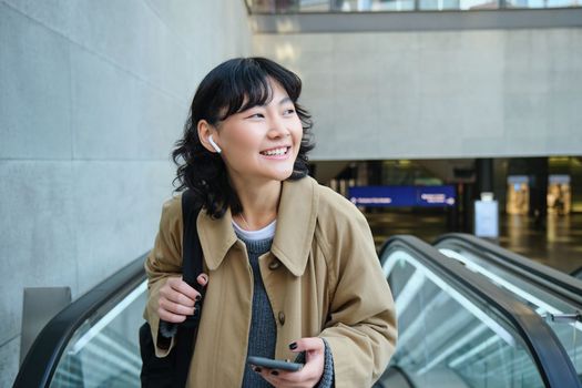 Portrait of brunette girl in wireless headphones, listens music, uses smartphone, goes on escalator. Copy space