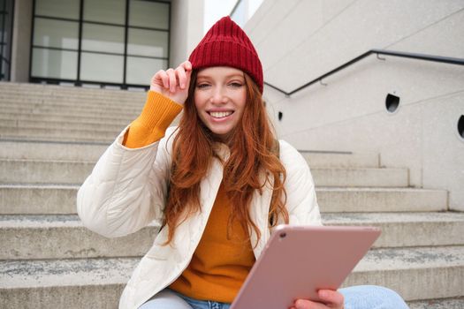 Beautiful redhead female model, ginger girl with digital tablet, sits on stairs outdoors, reads on her gadget, uses internet application and wifi on street.