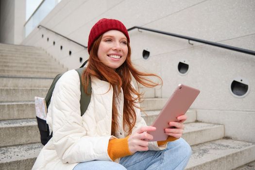 Young stylish girl, redhead female students sits on stairs outdoors with digital tablet, reads, uses social media app on gadget, plays games while waits on street.