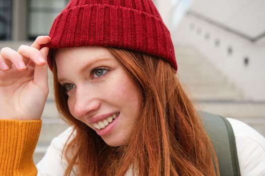 Stylish redhead girl in warm red hat, smiling relaxed, sitting with backpack on stairs near building, waits for someone outdoors.